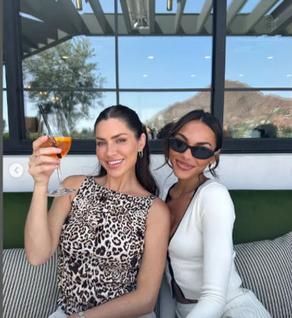 Two friends smiling on a sunny rooftop patio lounge with a mountain view through large glass windows, one raising an orange cocktail for a celebratory brunch.