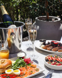 Sunny outdoor patio brunch: chilled champagne in an ice bucket with two flutes, smoked salmon bagel with soft-boiled eggs and greens, berry-topped granola bowl and French toast with fresh berries.
