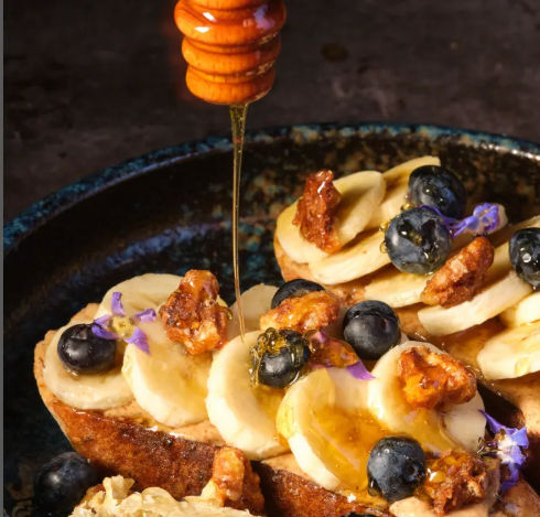 Close-up of gourmet brunch toast topped with banana slices, blueberries, candied walnuts and tiny purple edible flowers, golden honey drizzling from a wooden dipper.