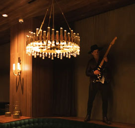 Dimly lit wood-paneled lounge with a sparkling crystal chandelier and a guitarist in a hat playing an electric guitar on a raised ledge.