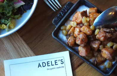 Cast-iron skillet of crispy golden cauliflower bites with grapes and nuts, nearby mixed greens salad and utensils on a wooden table at a Nashville restaurant.