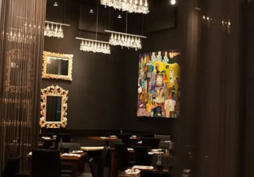 Ambient upscale restaurant dining room with dark walls, hanging wine-glass chandeliers, ornate gold-framed mirrors, vibrant modern artwork, and neatly set wooden tables and chairs.