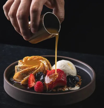 Hand drizzling warm caramel sauce over a plated gourmet dessert: mini bundt cake, fresh strawberries, raspberries, blackberries, vanilla ice cream and crumble on a dark plate.
