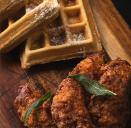 Crispy golden fried chicken pieces garnished with sage beside a powdered Belgian waffle on a wooden board — classic Southern-style chicken and waffles.