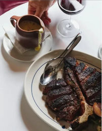Sliced charred bone-in steak with roasted garlic on an oval plate, creamy mustard sauce being poured from a pitcher, spoon and glass of red wine on a white table