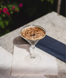 Espresso martini in a chilled coupe glass topped with cocoa dusting and three coffee beans, set on a marble table with a navy napkin and blurred garden background