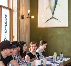 Group of diners sitting at a restaurant bar, chatting over wine and cocktails beneath warm wall sconces and a large framed fish painting on an olive-green wall.