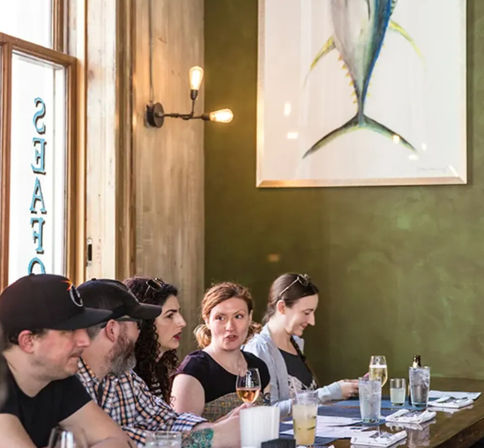 Group of diners sitting at a restaurant bar, chatting over wine and cocktails beneath warm wall sconces and a large framed fish painting on an olive-green wall.