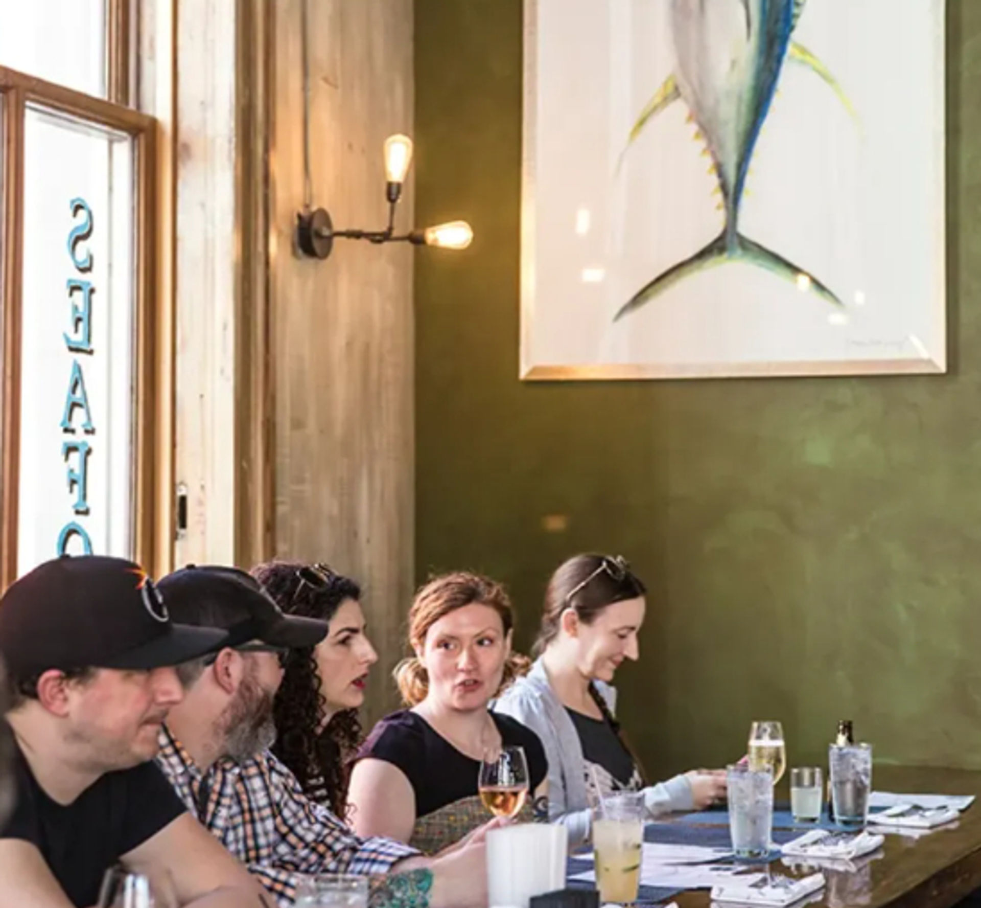 Group of diners sitting at a restaurant bar, chatting over wine and cocktails beneath warm wall sconces and a large framed fish painting on an olive-green wall.