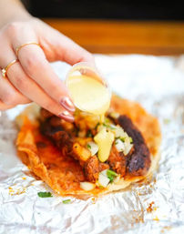 Hand pouring creamy yellow sauce from a small plastic cup onto a street-style taco with grilled meat, chopped onions and cilantro on a crispy tortilla resting on foil