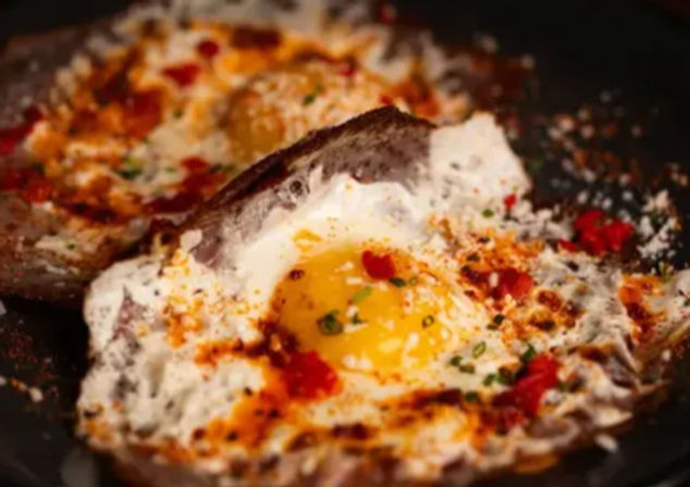 Two fried-egg-in-toast slices in a skillet with runny golden yolks, melted cheese, red chili flakes and chopped chives — savory spicy breakfast toast.