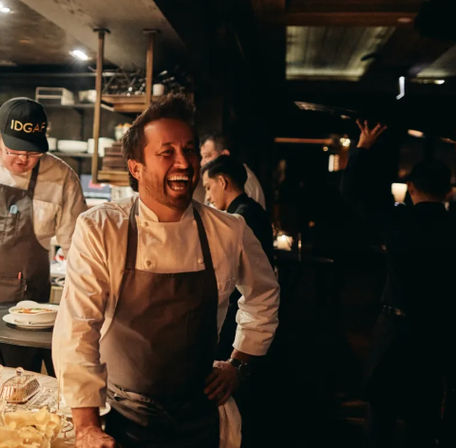 Smiling chef in white coat and apron laughing in a busy, dimly lit restaurant kitchen with staff working in the background