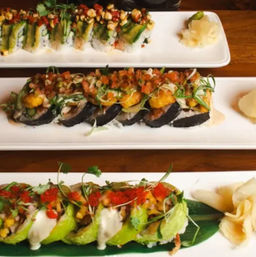 Vibrant assortment of gourmet sushi rolls on white rectangular plates over a wooden table, topped with avocado, spicy mayo, tobiko, microgreens and pickled ginger.