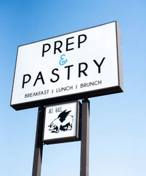 Roadside white rectangular café sign against a clear blue sky advertising breakfast, lunch and brunch; bold black lettering with a blue ampersand and a smaller panel below showing a winged pig illustration and the words “All Hail”.
