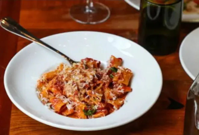 Hearty tomato pasta with grated Parmesan and parsley in a white bowl, fork on the rim, on a wooden table with a wine glass and bottle