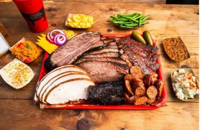 Colorful Texas-style BBQ platter on a wooden table with sliced brisket, smoked turkey, sausage links and burnt ends on a red tray, surrounded by pickles and sides like green beans, potato salad, coleslaw, mac and cheese and cornbread.