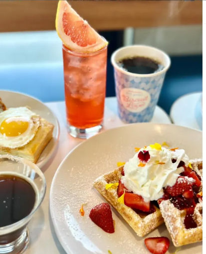 Brunch spread: strawberry-topped Belgian waffles with whipped cream and powdered sugar, a savory pastry with a fried egg, two cups of black coffee, and a tall pink grapefruit soda garnished with a grapefruit wedge.