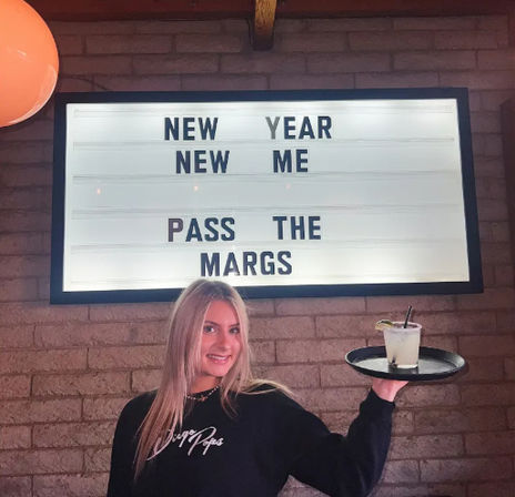 Smiling person holding a tray with a margarita in front of a backlit marquee sign on a brick wall that reads "NEW YEAR NEW ME PASS THE MARGS" in a lively bar setting.