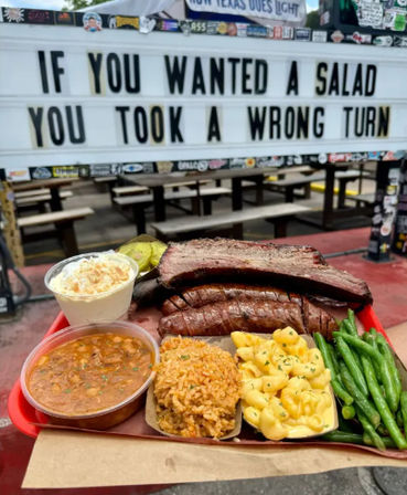 Heaping BBQ tray with smoked brisket, sausage, mac & cheese, rice, green beans, beans, slaw and pickles in front of an outdoor picnic-style patio and a marquee sign reading “If you wanted a salad you took a wrong turn.”