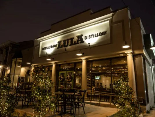 Cozy nighttime restaurant and distillery storefront with warm lights, outdoor patio seating, string lights and potted greenery on the sidewalk.