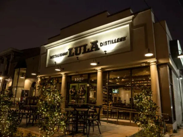 Cozy nighttime restaurant and distillery storefront with warm lights, outdoor patio seating, string lights and potted greenery on the sidewalk.