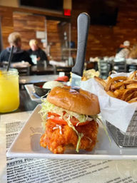 Crispy fried chicken sandwich with shredded lettuce and tomato on a brioche bun, knife stuck through it, served with curly fries in a metal basket and a mason-jar drink on a rustic casual restaurant bar table with wood-paneled interior.