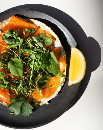 Overhead brunch shot of an open-faced smoked salmon bagel with cream cheese, fresh microgreens and herbs, served with a lemon wedge on a black plate.
