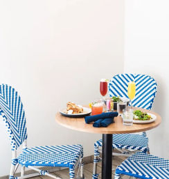 Bright brunch scene: small round wooden table with mimosas, cocktails, salad and pastry, set with navy napkins and surrounded by blue-and-white woven bistro chairs against a white wall.