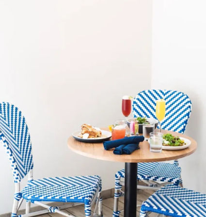 Bright brunch scene: small round wooden table with mimosas, cocktails, salad and pastry, set with navy napkins and surrounded by blue-and-white woven bistro chairs against a white wall.