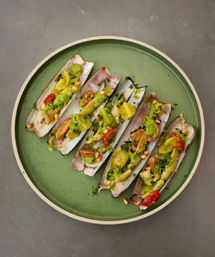 Mediterranean-style razor clams on a green plate topped with herb pesto, cherry tomatoes, parsley and pine nuts — grilled seafood appetizer