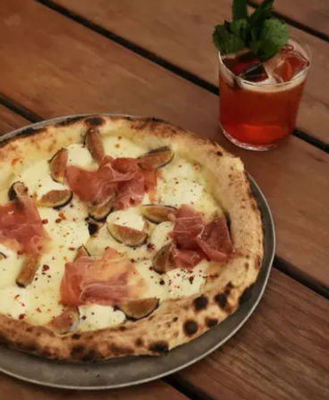 Neapolitan-style pizza with melted mozzarella, fig slices and prosciutto on a charred crust on a metal plate atop a rustic wooden table, paired with a red cocktail garnished with mint.