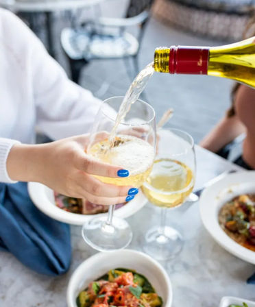 Hand with bright blue nail polish holding a wine glass as white wine is poured at an outdoor bistro table with bowls of salad and pasta.