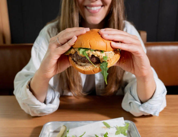 Close-up of a smiling person holding a juicy gourmet burger with arugula and creamy sauce in a brioche bun at a casual restaurant table.