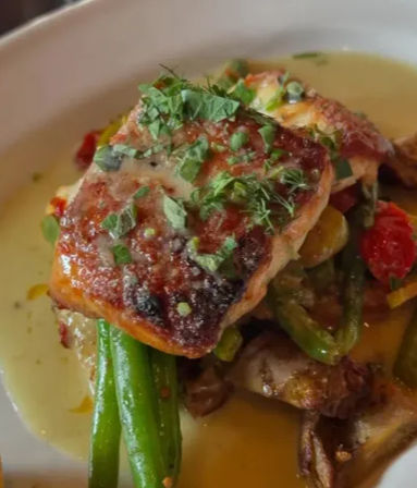 Seared salmon fillet topped with chopped herbs, served over crisp green beans, cherry tomatoes and sautéed mushrooms in a creamy lemon-butter sauce on a white plate.