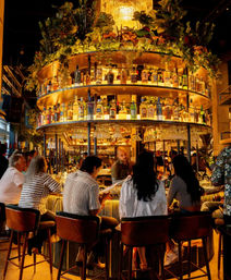 Group of patrons seated around a circular, backlit cocktail bar with tiers of liquor bottles, hanging plants and warm chandelier lighting — lively upscale indoor bar scene at night
