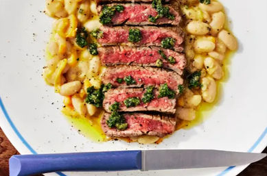 Top-down close-up of sliced medium-rare steak drizzled with green herb sauce on a bed of white beans, served on a white plate with a blue rim and a blue-handled knife — colorful hearty dinner plate
