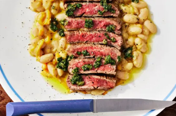 Top-down close-up of sliced medium-rare steak drizzled with green herb sauce on a bed of white beans, served on a white plate with a blue rim and a blue-handled knife — colorful hearty dinner plate