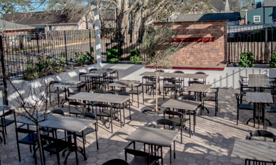 Sunny outdoor dining patio in an urban courtyard with rows of black metal tables and chairs on hexagonal pavers, small trees and a brick feature wall.