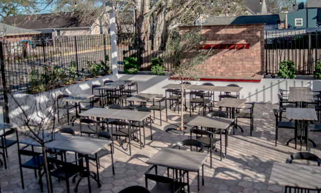 Sunny outdoor dining patio in an urban courtyard with rows of black metal tables and chairs on hexagonal pavers, small trees and a brick feature wall.