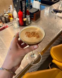Hand holding a coupe glass with a creamy cocktail topped by branded foam reading "Milk & Honey" on a stainless bar counter with condiments and yellow barstools