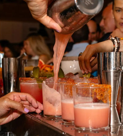 Bartender pouring frothy pink cocktails into a row of short glasses rimmed with chili-salt and garnished with lime at a busy bar