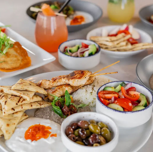Colorful Mediterranean mezze platter with grilled chicken skewers over rice, warm pita triangles, mixed olives, cucumber–tomato salad and creamy dips, cocktails blurred in background