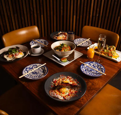 Asian-fusion brunch spread on a warm wood restaurant table: central noodle bowl, fried-egg plates with chili oil and herbs, small appetizers on rectangular plates, blue-and-white patterned side plates with chopsticks, coffee, orange juice and a champagne flute.