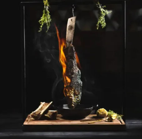 Dramatic food photo of a flaming charred beef rib suspended from a hook over a cast-iron pan on a wooden board with roasted bone, lemon and fresh herbs — modern fine-dining smoked meat presentation.