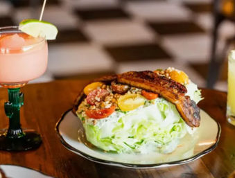 Iceberg wedge salad with cherry tomatoes, crumbled nuts and caramelized fried plantains on a plate, served beside a pink cocktail and yellow drink on a wooden table in a casual restaurant with a checkered floor.