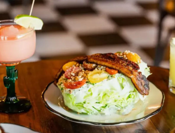 Iceberg wedge salad with cherry tomatoes, crumbled nuts and caramelized fried plantains on a plate, served beside a pink cocktail and yellow drink on a wooden table in a casual restaurant with a checkered floor.