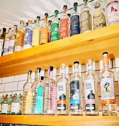 Bright wooden bar shelves lined with colorful mezcal and tequila bottles against a white subway-tile backsplash, neatly arranged and ready for service.