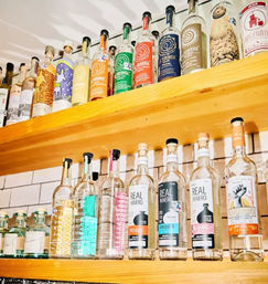 Bright wooden bar shelves lined with colorful mezcal and tequila bottles against a white subway-tile backsplash, neatly arranged and ready for service.