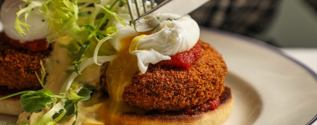 Brunch plate with a crispy breaded croquette on a toasted English muffin, topped by a runny poached egg, tomato relish and frisée greens.