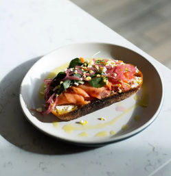 Gourmet smoked salmon toast on rustic sourdough topped with pickled red onions, microgreens and crumbled egg, olive oil drizzle on a white plate on a marble countertop — brunch dish
